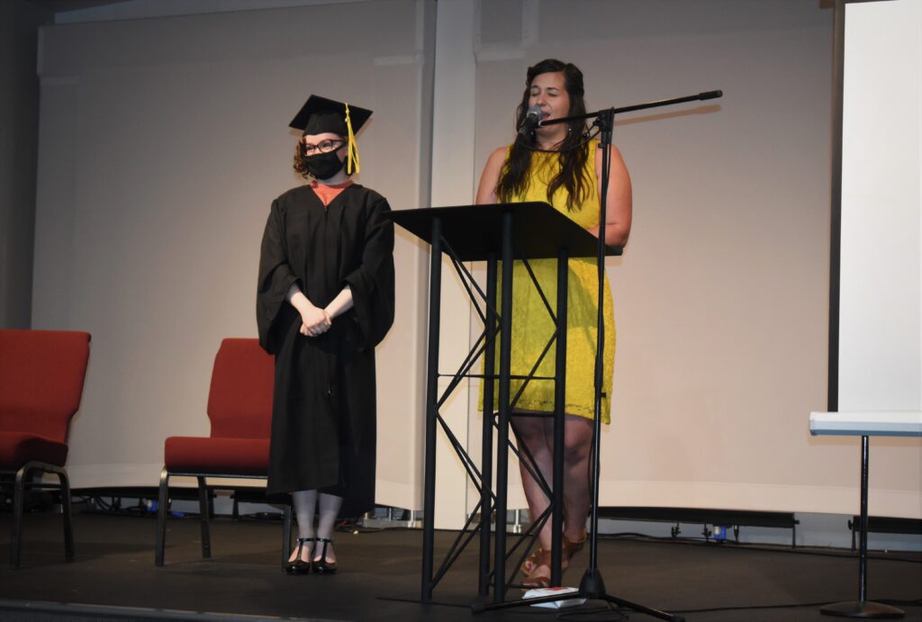 Maggie Sanazaro, a tall, dark haired woman wearing a yellow dress, stands at a podium. On her left is Arianna Brooks, a shorter teen girl with curly hair and glasses. She is wearing heels, a graduation gown, and a graduation cap. 